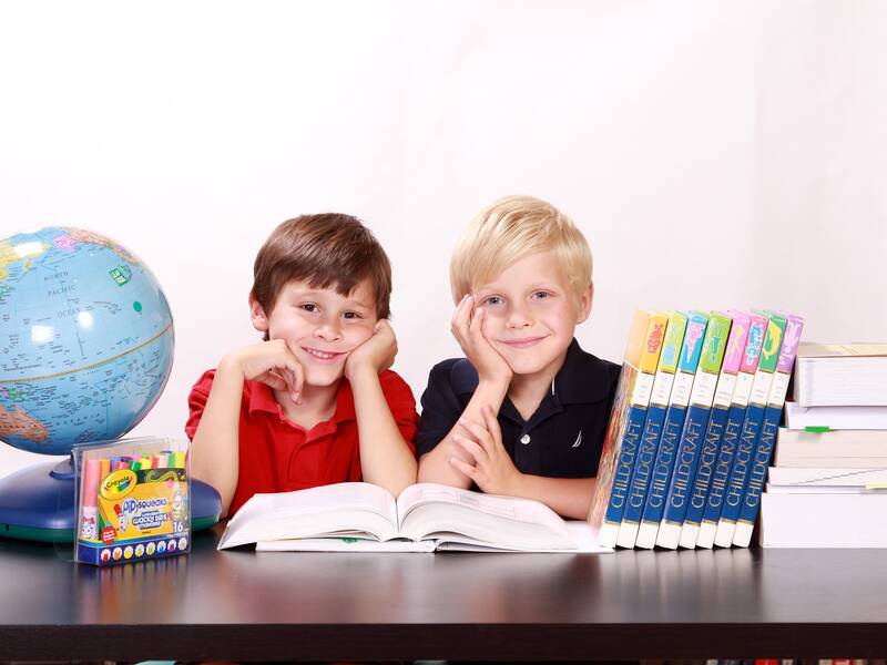 Zwei lächelnde Kinder sitzen an einem Tisch, auf dem Schulbücher, ein Globus und Buntstifte liegen