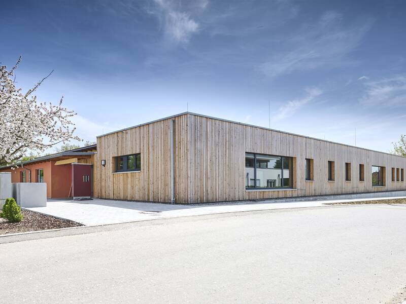 Außenansicht des Kindergartens "Löwenzahn" mit Holzverkleidung unter blauem Himmel, daneben blühender Baum