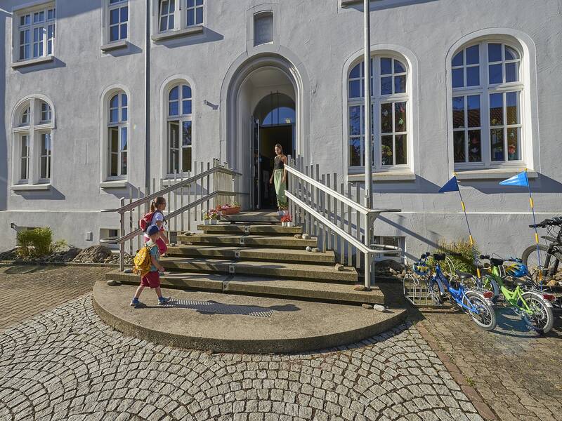 Haupteingang des Kindergartens in Grüningen, Erzieherin nimmt ankommende Kinder in Empfang