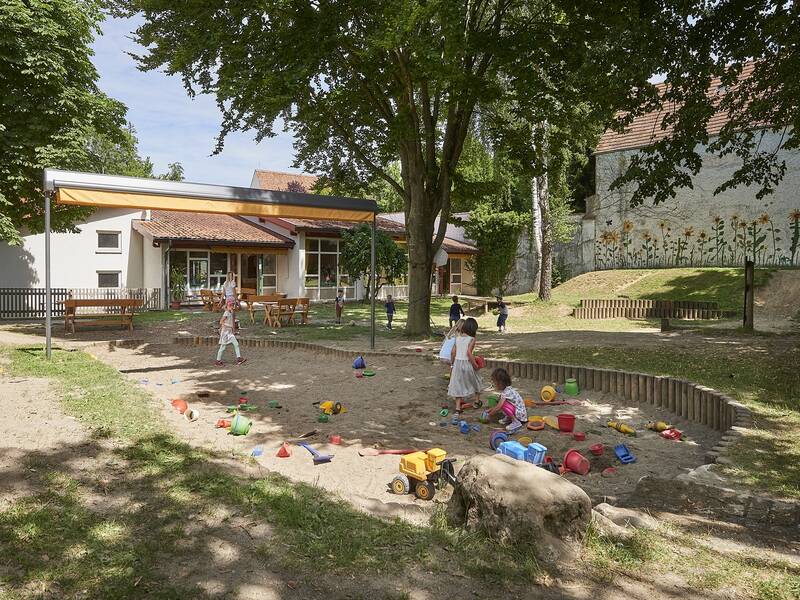 Sandkasten des Kindergartens Storchennest mit Spielzeug und spielenden Kindern, umgeben von einer Grünfläche