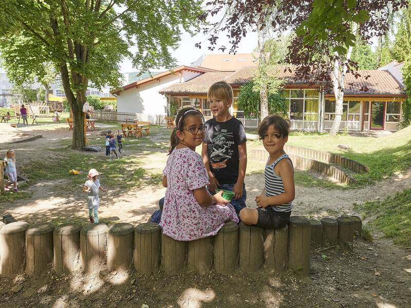 Drei Kinder vor dem Hintergrund des Außengeländes des Kindergartens Storchennest mit Spielgeräten und spielenden Kindern