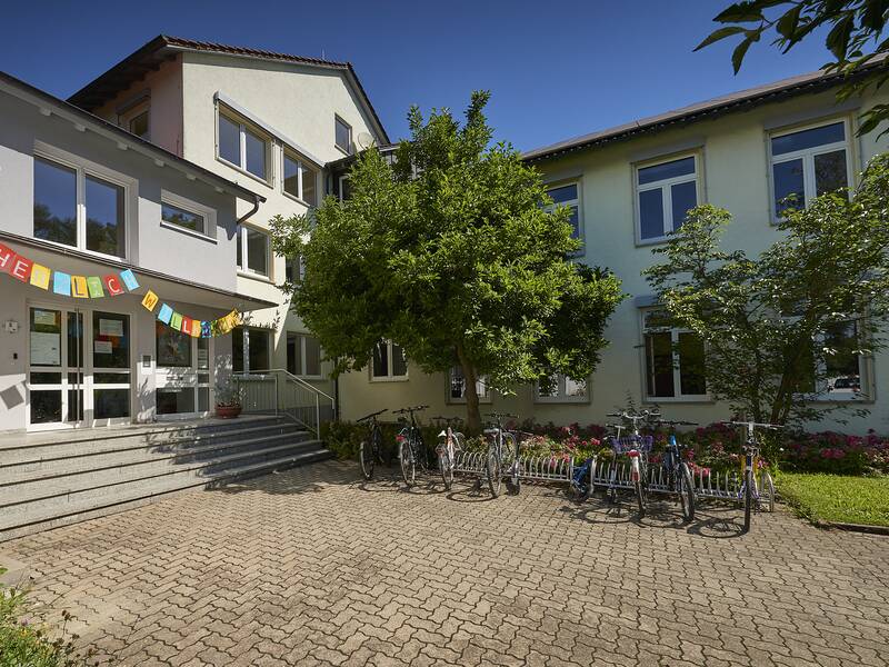 Ein sonniger Blick auf das zweistöckige Schulgebäude der Sankt Gerhard-Schule mit einer Reihe geparkter Fahrräder vorne und einem Banner über dem Eingang, der "HERZLICH WILLKOMMEN" sagt.